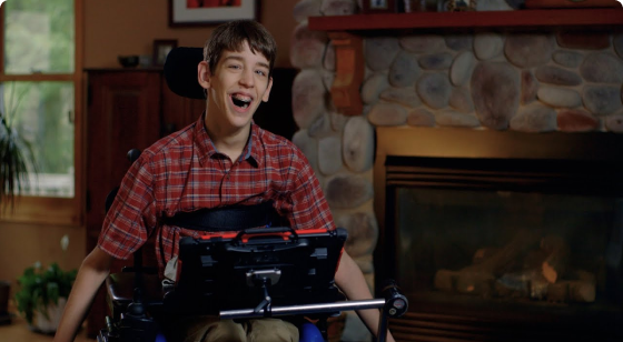 boy in wheelchair with laptop attached; he’s smiling and there’s a large fireplace in the background